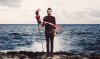 A Man is standing in Front of the Ocean carrying a Stick and wearing a red shirt with blue Jeans Trousers. In the background is the sky.